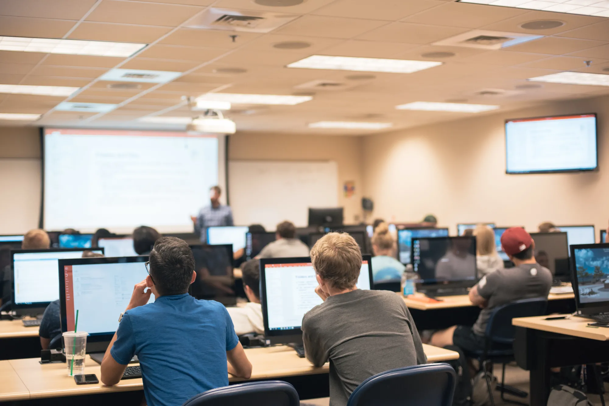 Students in a classroom listen to a lecture while working on computers. 